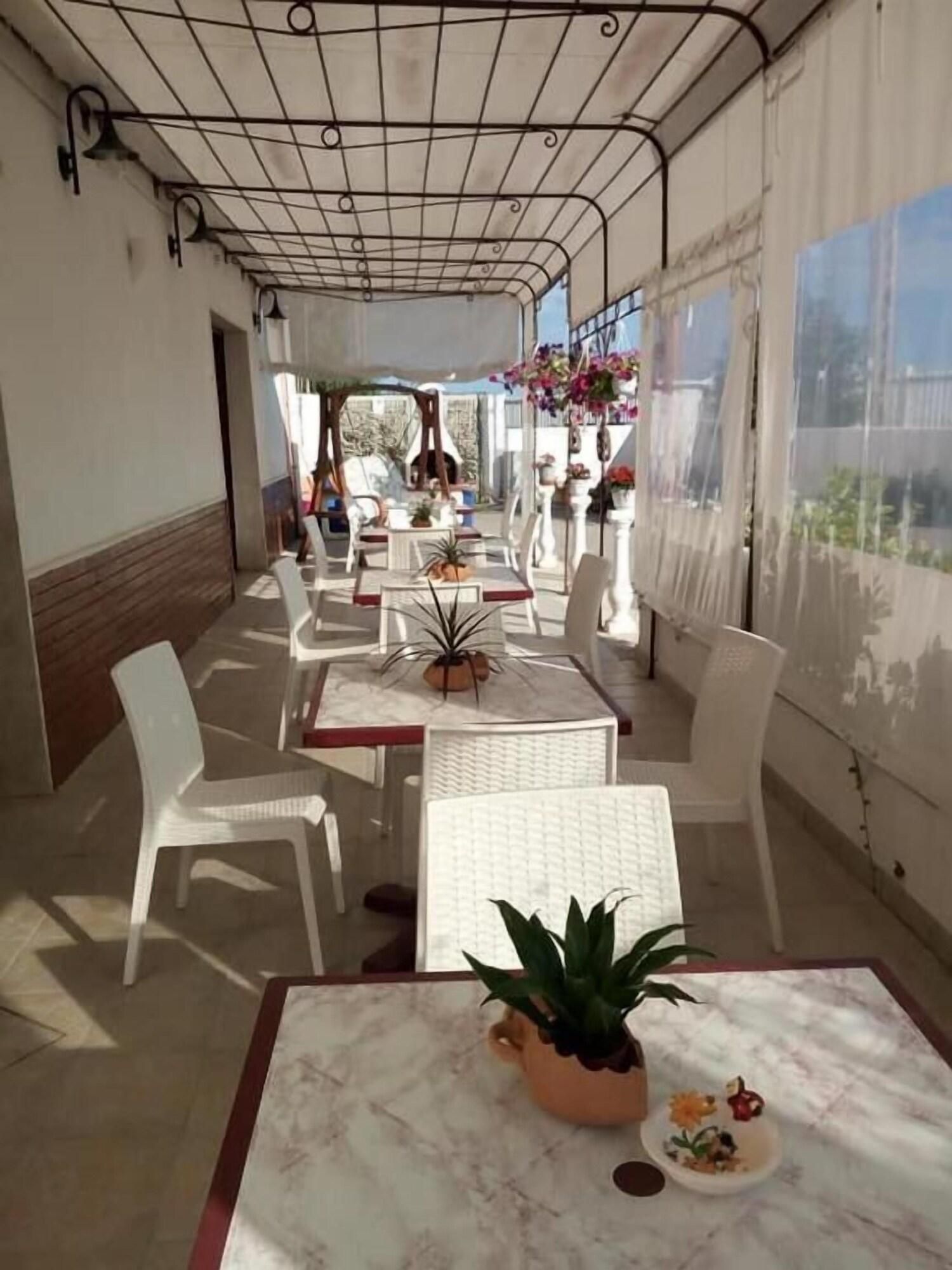 Un patio al aire libre con mesas y sillas blancas, plantas en macetas y cestas de flores colgantes.