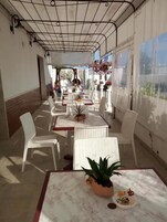 Un patio al aire libre con mesas y sillas blancas, plantas en macetas y cestas de flores colgantes.