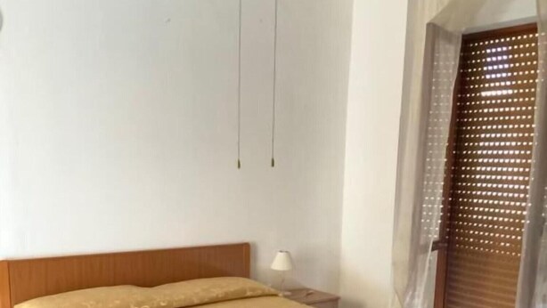 Ein ordentlich bezogenes Bett mit einem Holzkopfstück, ein Nachttisch mit einer Lampe und ein Fenster mit einer Holzjalousie.