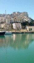 Un puerto deportivo con barcos amarrados, un edificio de fachada blanca y una colina con varias construcciones.