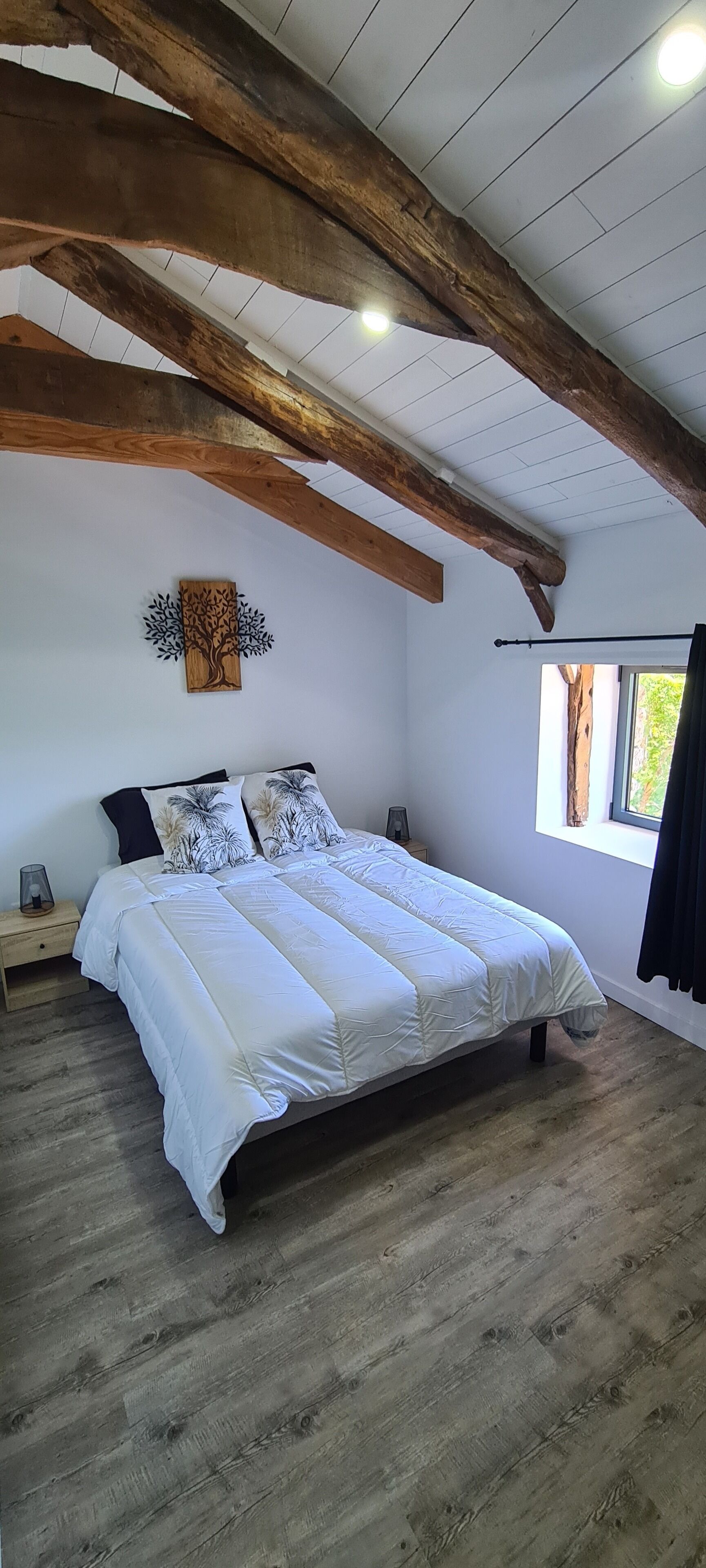 A bedroom with a wooden ceiling, a bed with white bedding, a wall-mounted tree decoration, and a window with a view of greenery.