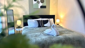 Ein ordentlich gemachtes Bett mit grauer Decke, zwei Kissen und einem auf dem Bett gefalteten weißen Handtuch. Auf jeder Seite des Bettes stehen zwei Nachttischlampen.