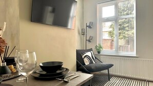 Ein modernes Wohnzimmer mit Esstisch, schwarz-weißen Kissen, einer schwarzen Couch und einem großen Fenster mit Jalousien.