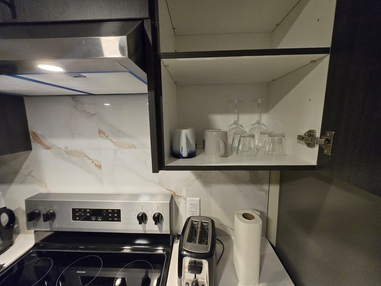A kitchen with a stove, toaster, and a shelf with glasses and a kettle.