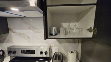 A kitchen with a stove, toaster, and a shelf with glasses and a kettle.