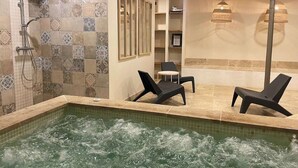 Een wellnessruimte met een jacuzzi, twee lounge fauteuils, een douchegedeelte en een betegelde muur met een decoratief patroon.