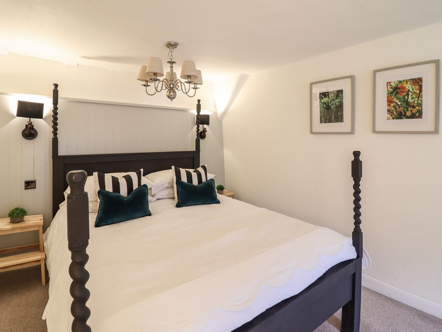 A four-poster bed with a white bedspread and teal pillows, a chandelier, and two framed pictures on the wall.