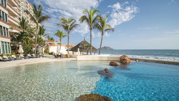 A clear pool with rocks, palm trees, and a beach view.