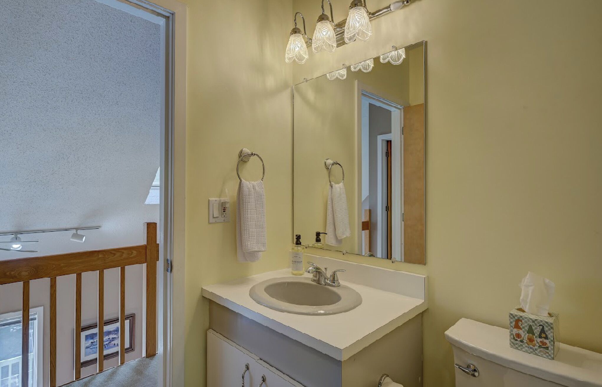 A bathroom with a white sink, a mirror, and a toilet. There is a towel hanging on the wall and a tissue box on the toilet.