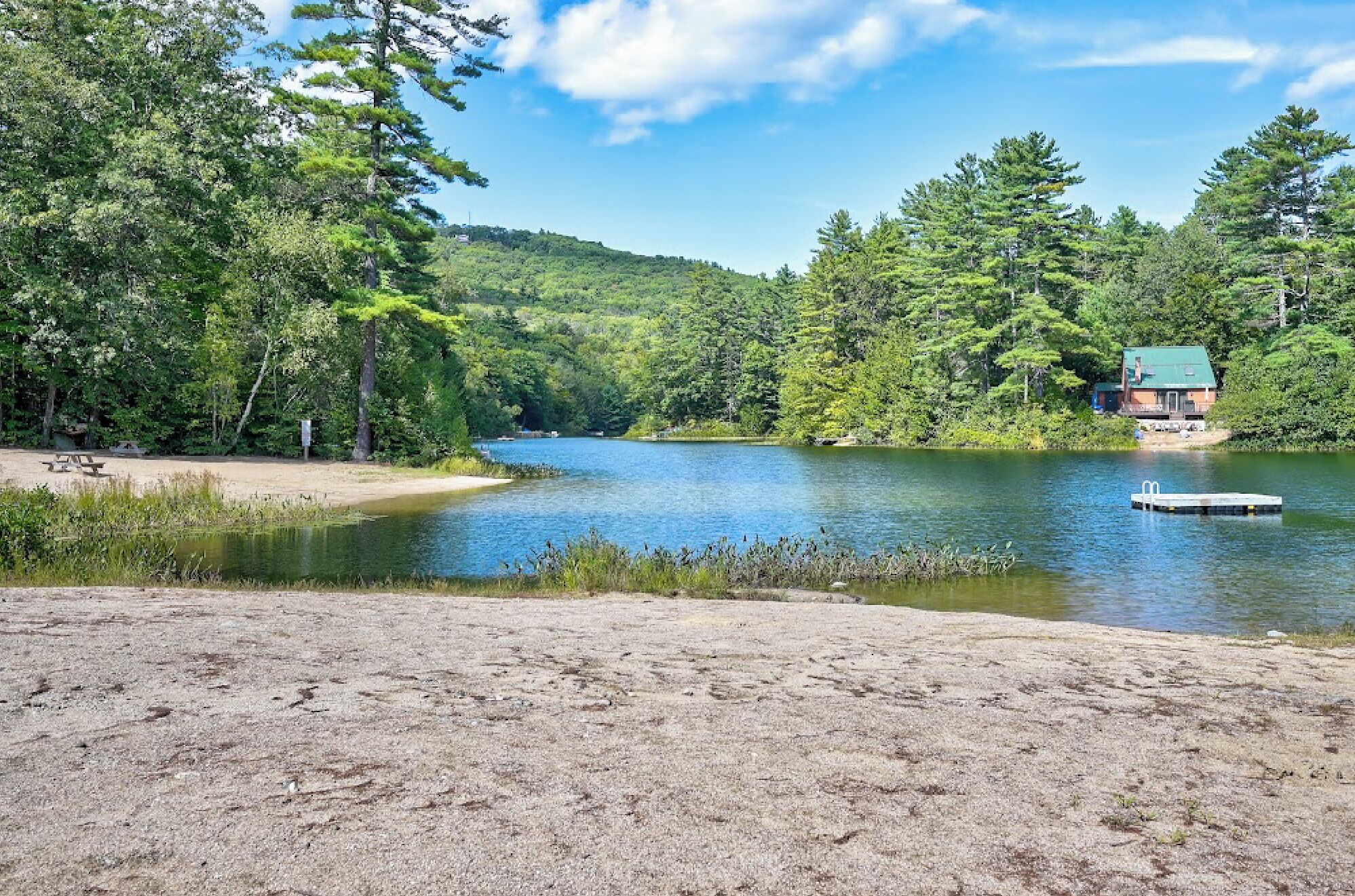 Uma cabana à beira do lago com um cais, rodeada por uma floresta densa e um céu claro.