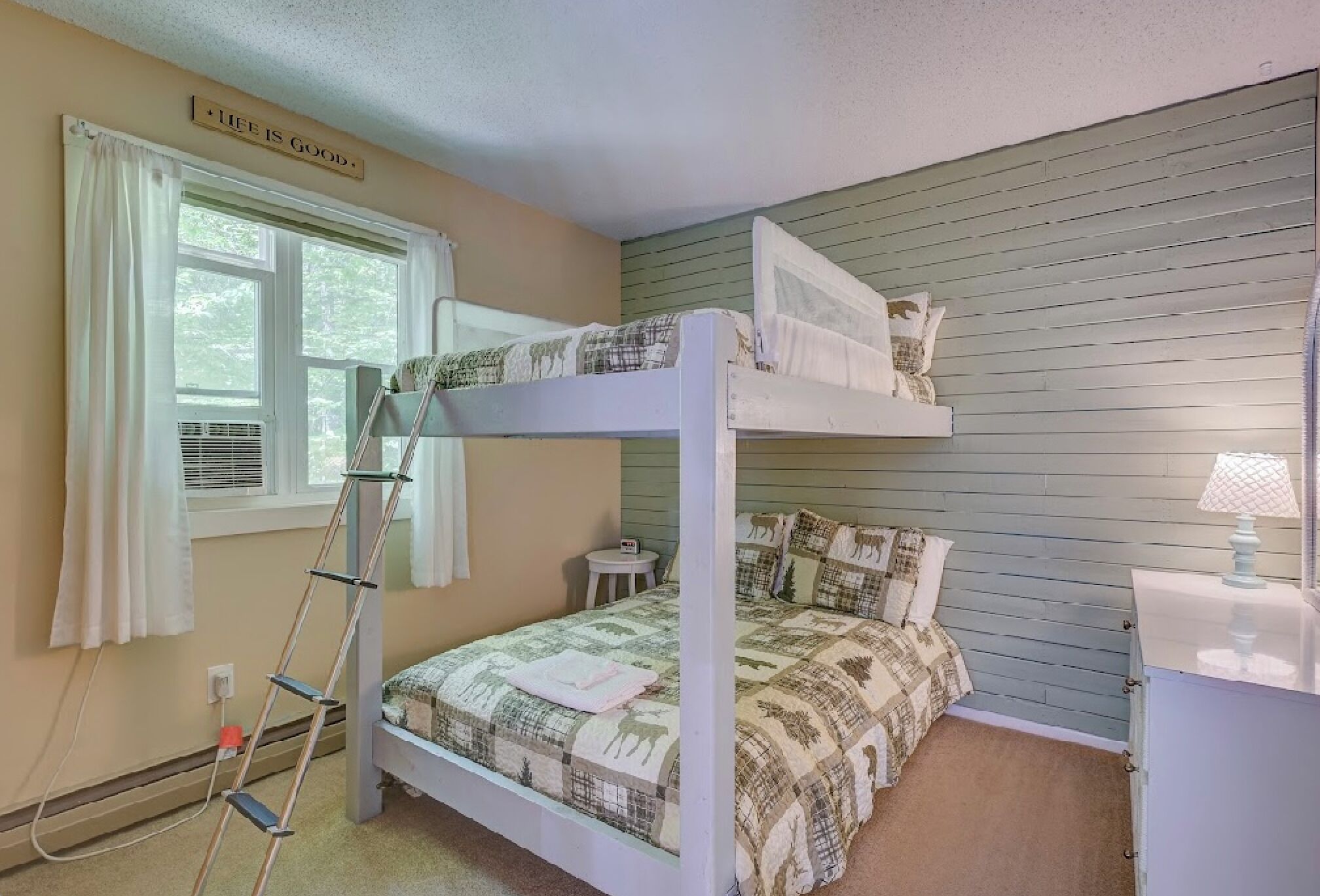 A bunk bed room with a ladder, a small bedside table, and a window with curtains.