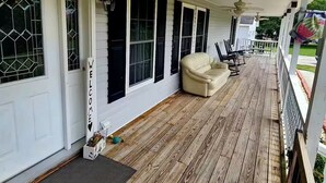 Un porche abrité avec un plancher en bois, un canapé beige et une porte blanche ornée d’une pancarte « BIENVENUE ».