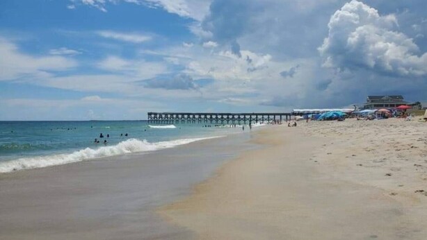 Une plage avec une jetée qui s’avance dans l’océan, un rivage sablonneux, ainsi que quelques parasols et des personnes qui se prélassent sur la plage.