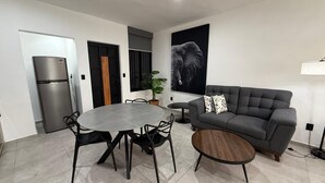 Uma sala de estar moderna com um sofá cinzento, uma mesa de jantar redonda com cadeiras, uma pequena mesa de centro de madeira, uma geladeira e um quadro grande na parede com a imagem de um elefante.