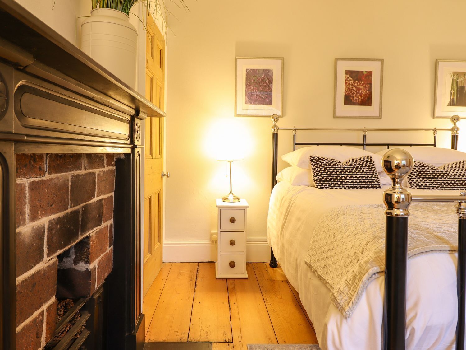 Une chambre avec une cheminée en briques, un lit agrémenté d’oreillers à motifs, une table de chevet avec une lampe et des tableaux encadrés accrochés au mur.