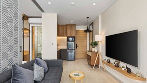 Een moderne woonkamer met een grijze bank, een kleine ronde tafel, een flatscreen televisie en een keuken op de achtergrond.