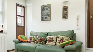 Ein grünes Sofa mit floralen Kissen, eine Wanduhr und motivierende Wandbilder in einem Raum mit weißen Wänden und einer Holztür.