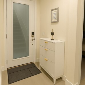A hallway with a white door, a small desk with drawers, a framed picture on the wall, and a doormat.
