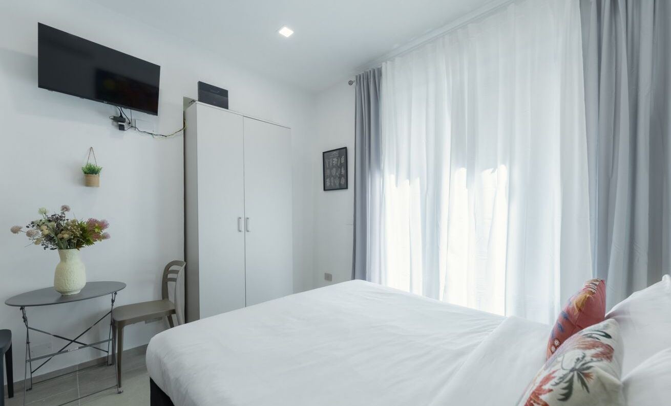 Ein Hotelzimmer mit Bett, Wandfernseher, einem kleinen Tisch mit Blumenvase und einem Kleiderschrank.
