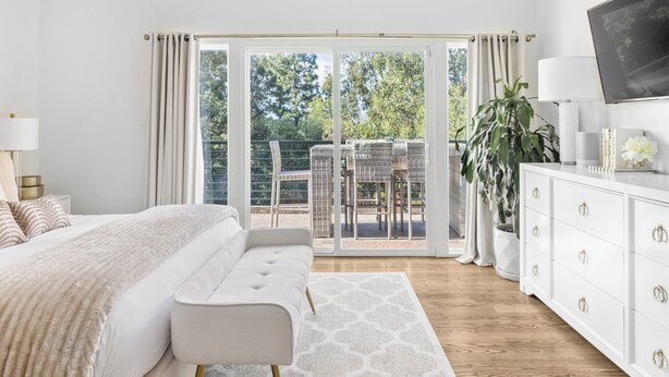 Une chambre à coucher comprenant un lit, un banc, une commode et donnant sur une terrasse extérieure avec des chaises et une table.