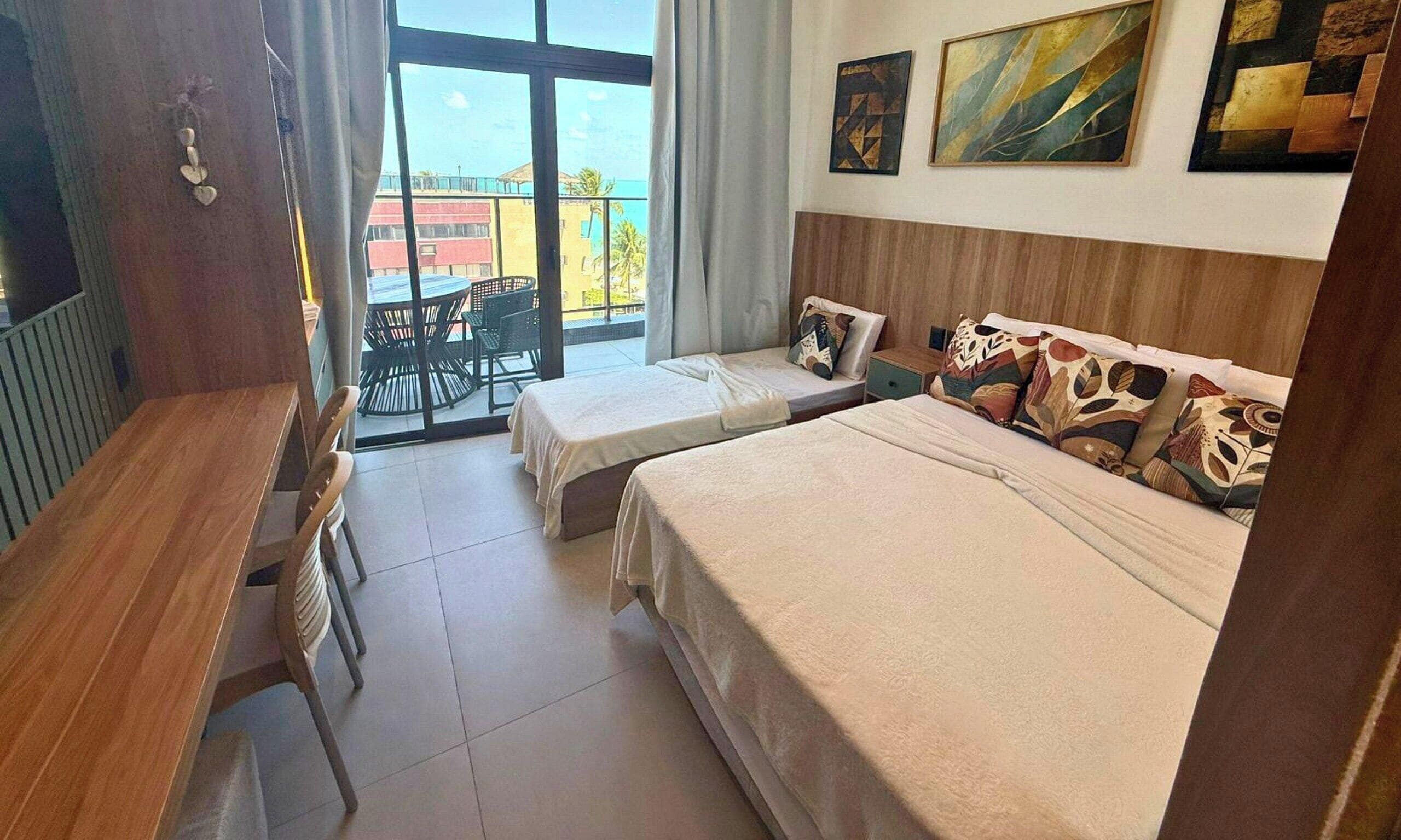 Habitación de hotel con dos camas, un escritorio de madera, un balcón con mobiliario exterior y obras de arte en las paredes.