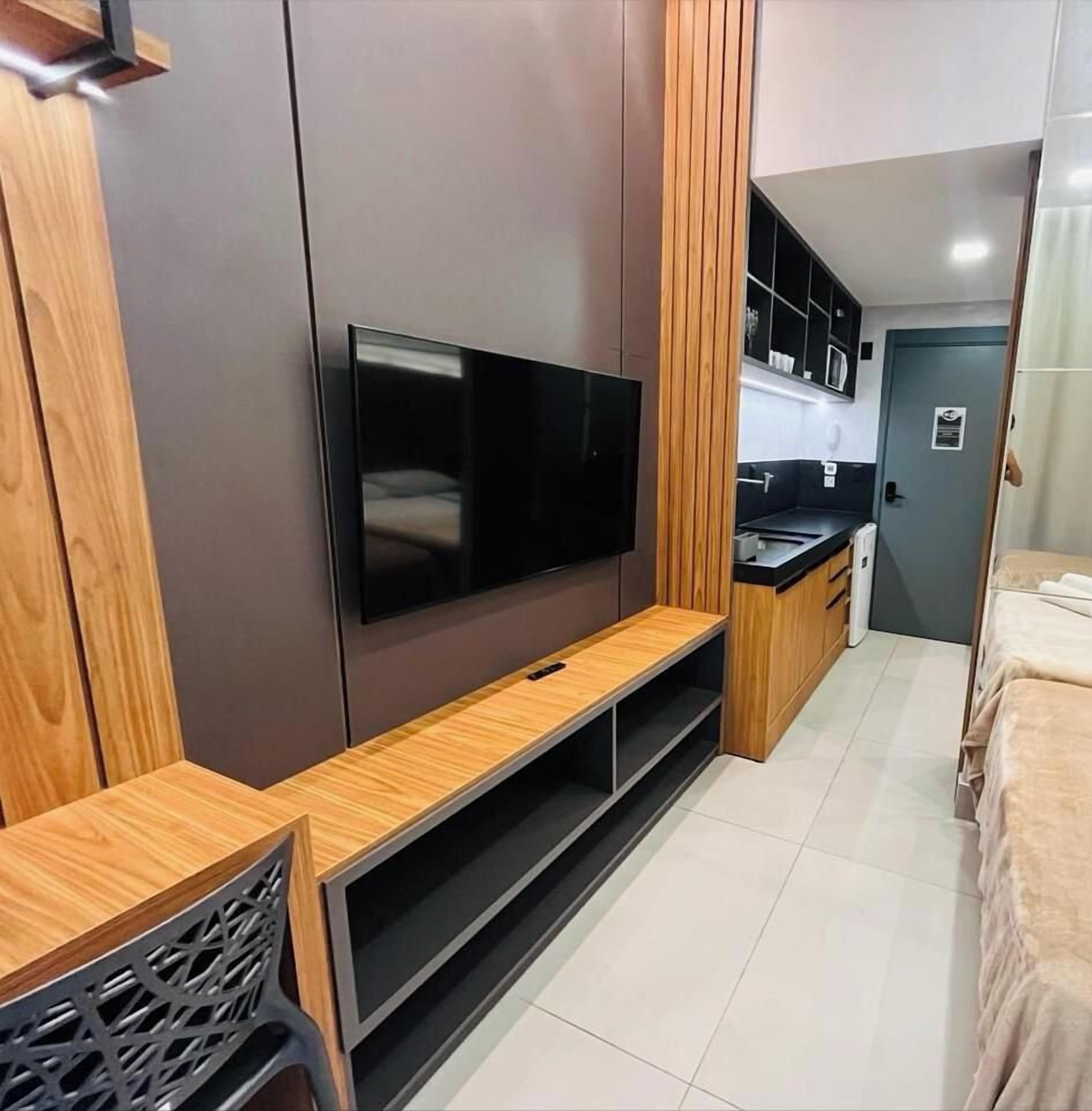 A compact room with a flat-screen TV mounted on a wall, a wooden bench, a kitchenette with cabinets, and a bed.