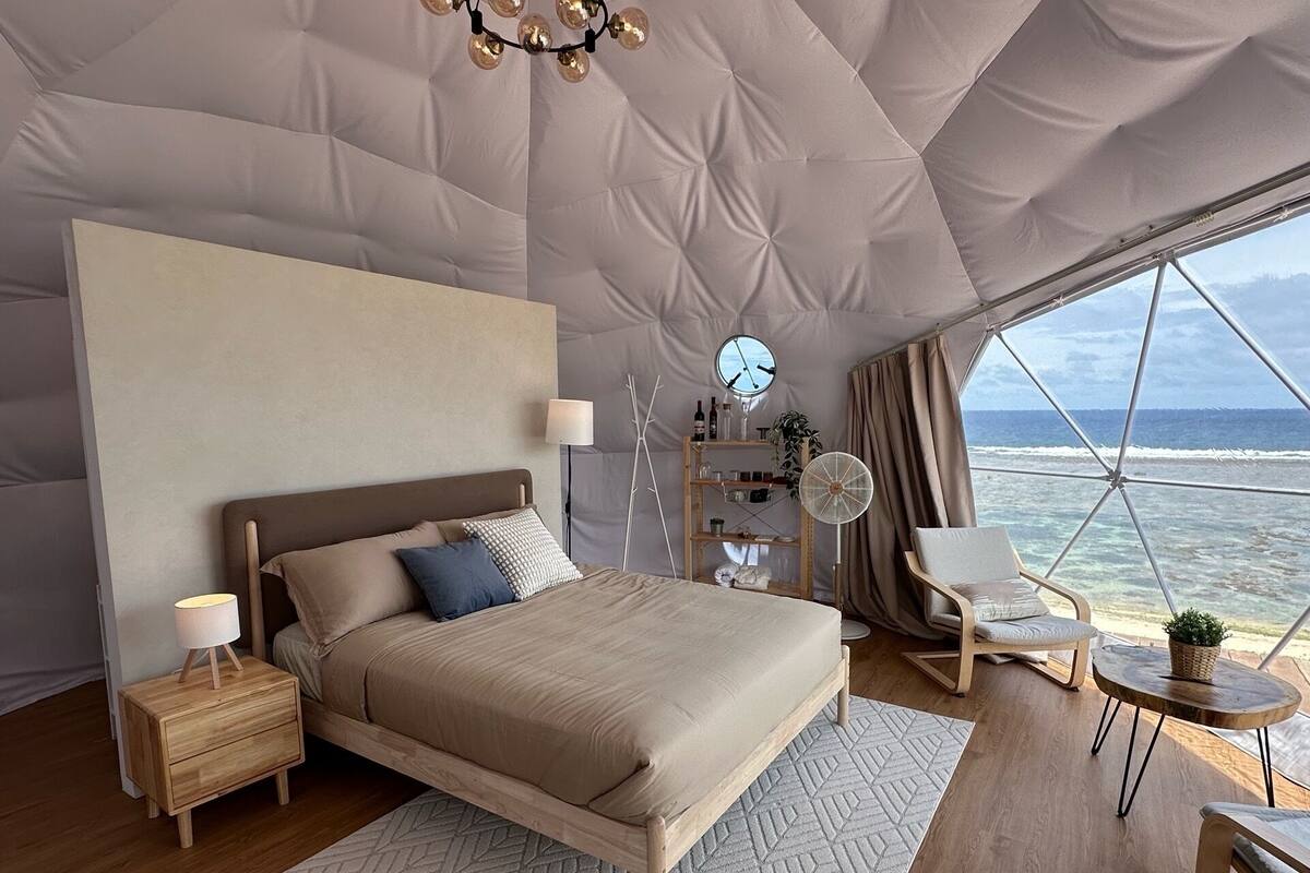 A bedroom with a bed, bedside tables, a nightstand, a chair, a small table, a fan, a clock, and a view of the ocean.