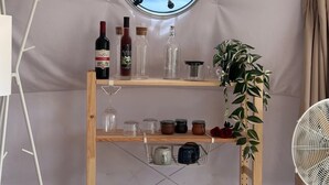 A room with a round window, wooden shelves with various items, a potted plant, and a fan. - Batanes Glamping (Sabtang)