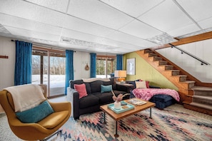 A cozy living room with a sofa, armchair, and coffee table, a staircase, and large windows with curtains.