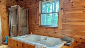 Interior de una cabaña de madera con una amplia bañera de madera, una ventana con persianas y un rústico armario de madera.