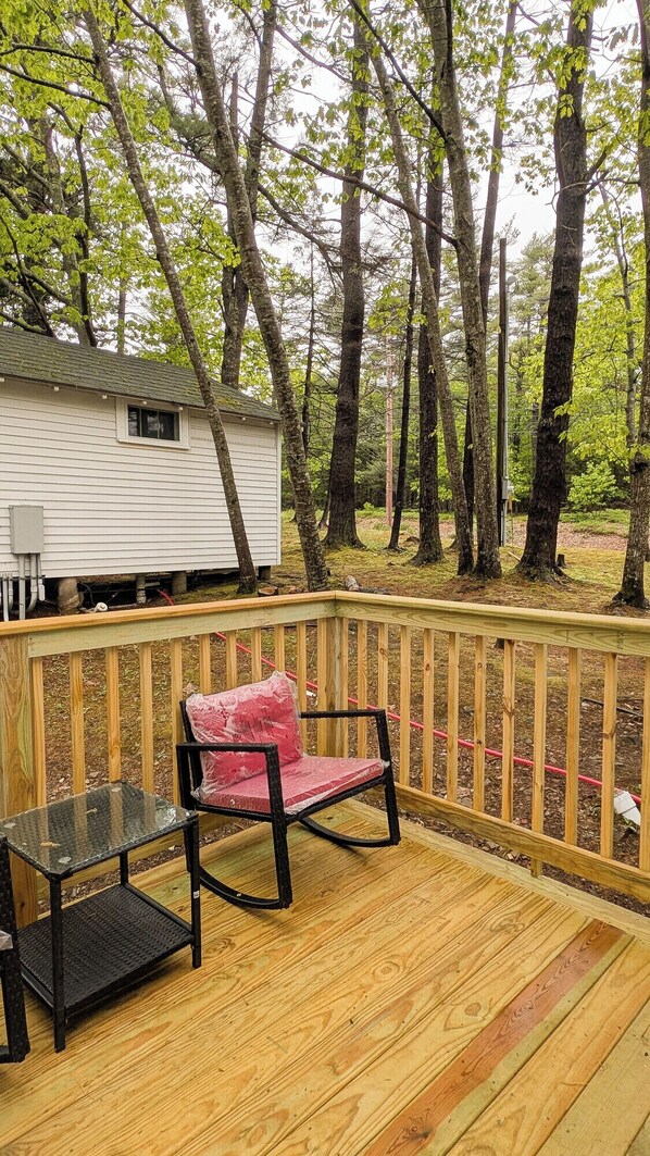 A wooden deck with a table and a chair, overlooking a forested area with a mobile home in the background.