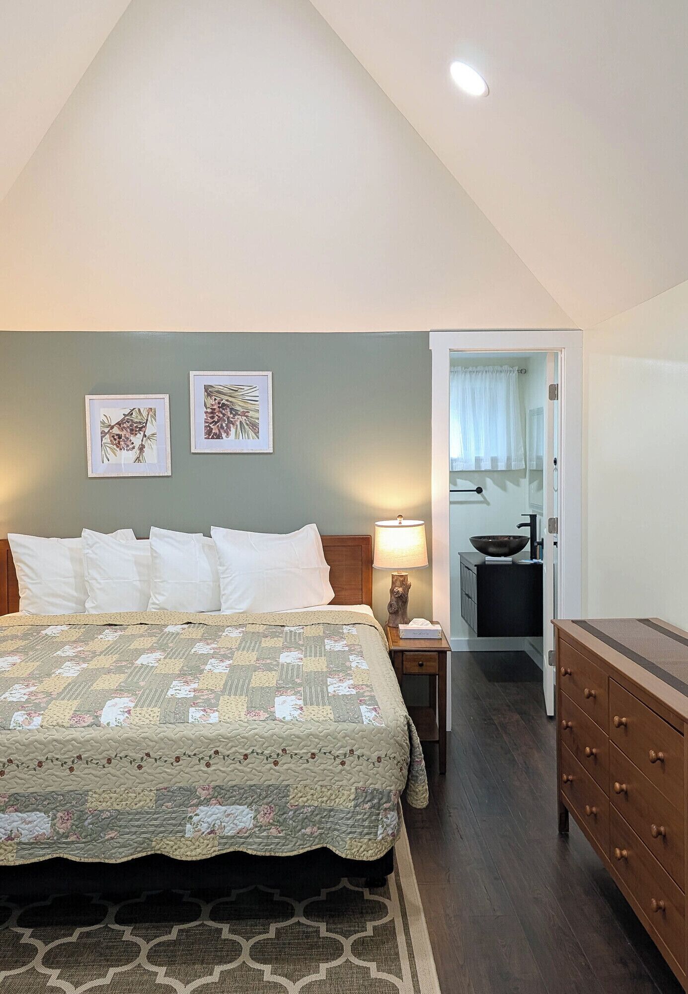 A bedroom with a bed, a nightstand, a dresser, and a bathroom visible through an open door.