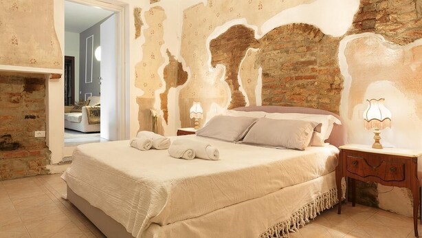A bedroom with a brick wall, a bed with a white bedspread, two bedside tables with lamps, and a door leading to another room. - One Bedroom Apartment (Vigevano)