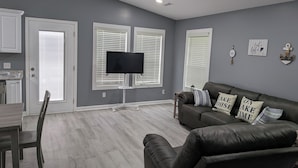 Een woonkamer met een donker leren bank, een flatscreen televisie op een standaard en een eethoek met een houten tafel en stoelen.
