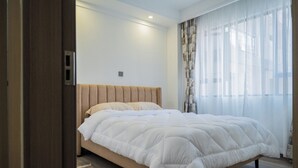 A bedroom with a bed, a wooden headboard, a window with sheer curtains, and a small rug on the floor. - Welcome to Summerville homes ,the cozy haven nestled in the heart of Nairobi. (Nairobi)