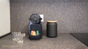A coffee maker with a black body and a wooden lid, accompanied by three clear glasses and a black thermos on a speckled countertop.