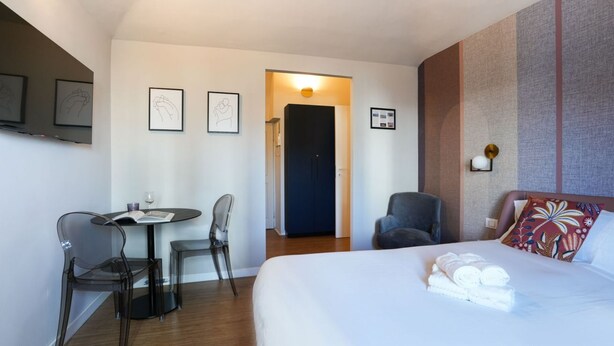 A hotel room with a bed, a desk with chairs, a door, and framed pictures on the wall.