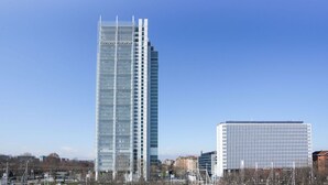 A tall, modern skyscraper with a glass facade and a shorter, white building with a grid-like pattern in the foreground.