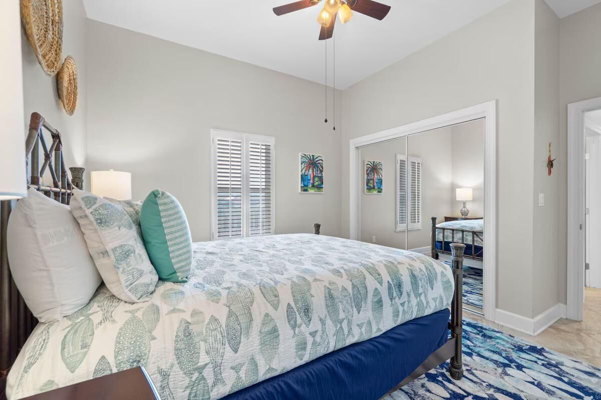Une chambre à coucher comprenant un lit, un ventilateur de plafond, une fenêtre avec des stores et une porte donnant sur une autre pièce.