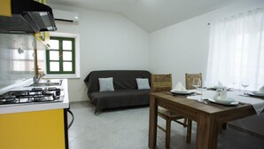 A compact kitchen with a gas stove, a dining table set for two, a sofa, and a window with curtains.