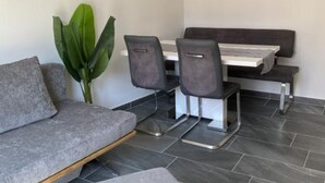 Una habitación moderna con un sofá gris, una mesa de comedor con cuatro sillas, una planta verde grande y un televisor de pantalla plana montado en la pared.