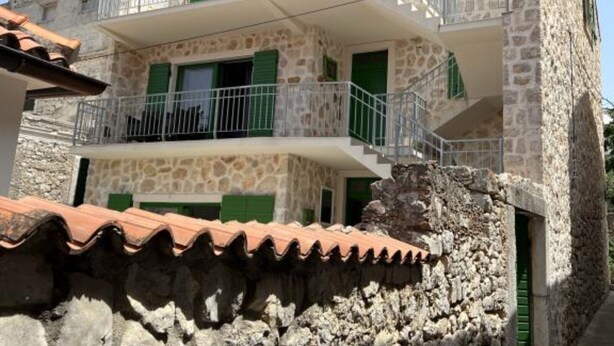 Un edificio de piedra de dos pisos con contraventanas verdes, un balcón y un muro de piedra al frente.
