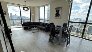 A spacious living area with a large window, a black sofa, a dining table, and a coffee table. - Luxe 2bed apartment with AC, fitness room in amazing Miami w/ beautiful sea view (Miami)