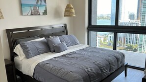 A modern bedroom with a large bed, a view of the city through large windows, and a beach scene artwork on the wall. - Luxe 2bed apartment with AC, fitness room in amazing Miami w/ beautiful sea view (Miami)