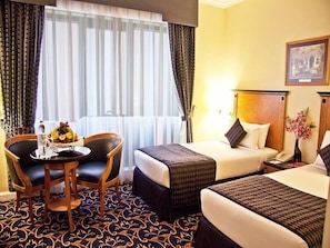 Quarto de hotel com duas camas, uma mesinha com uma cesta de frutas, uma cadeira e uma janela com cortinas.
