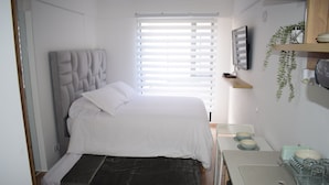 A bedroom with a bed, a bedside table with a lamp, a TV mounted on the wall, and a window with blinds.