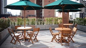 An outdoor patio with wooden tables and chairs, green umbrellas, and a cityscape view in the background.