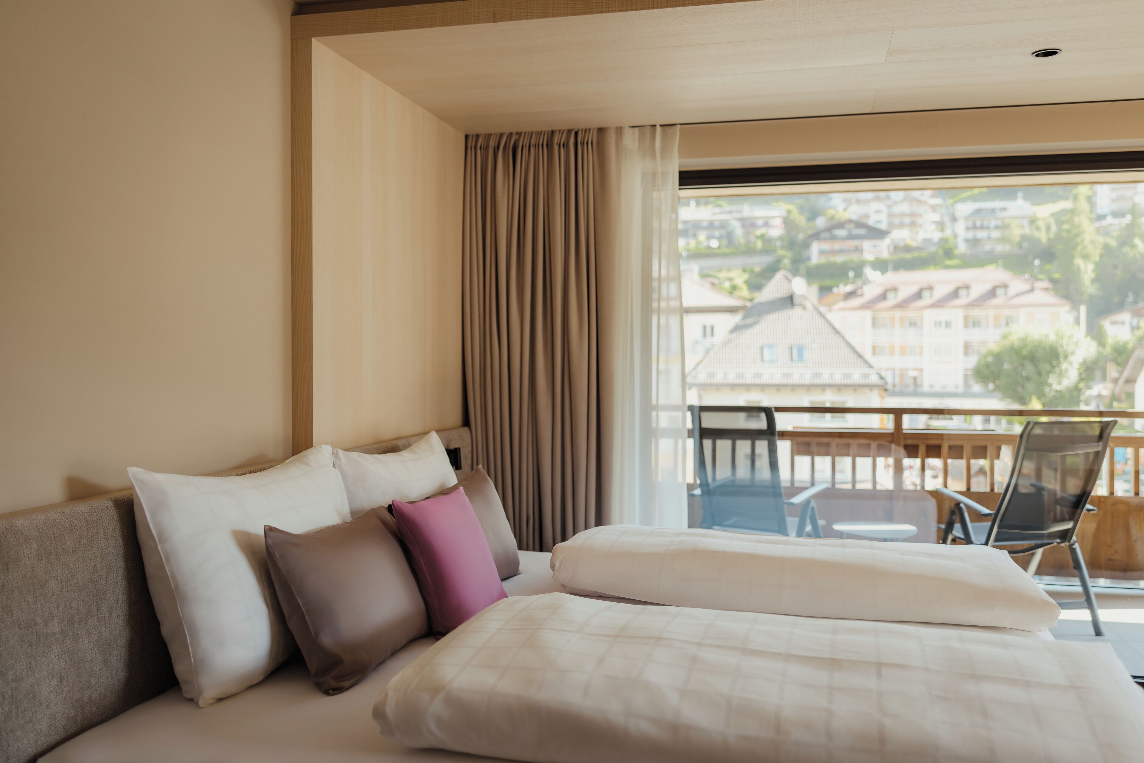 A hotel room with a bed, a view of buildings through a window, and a balcony with chairs.