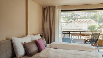 A hotel room with a bed, a view of buildings through a window, and a balcony with chairs.