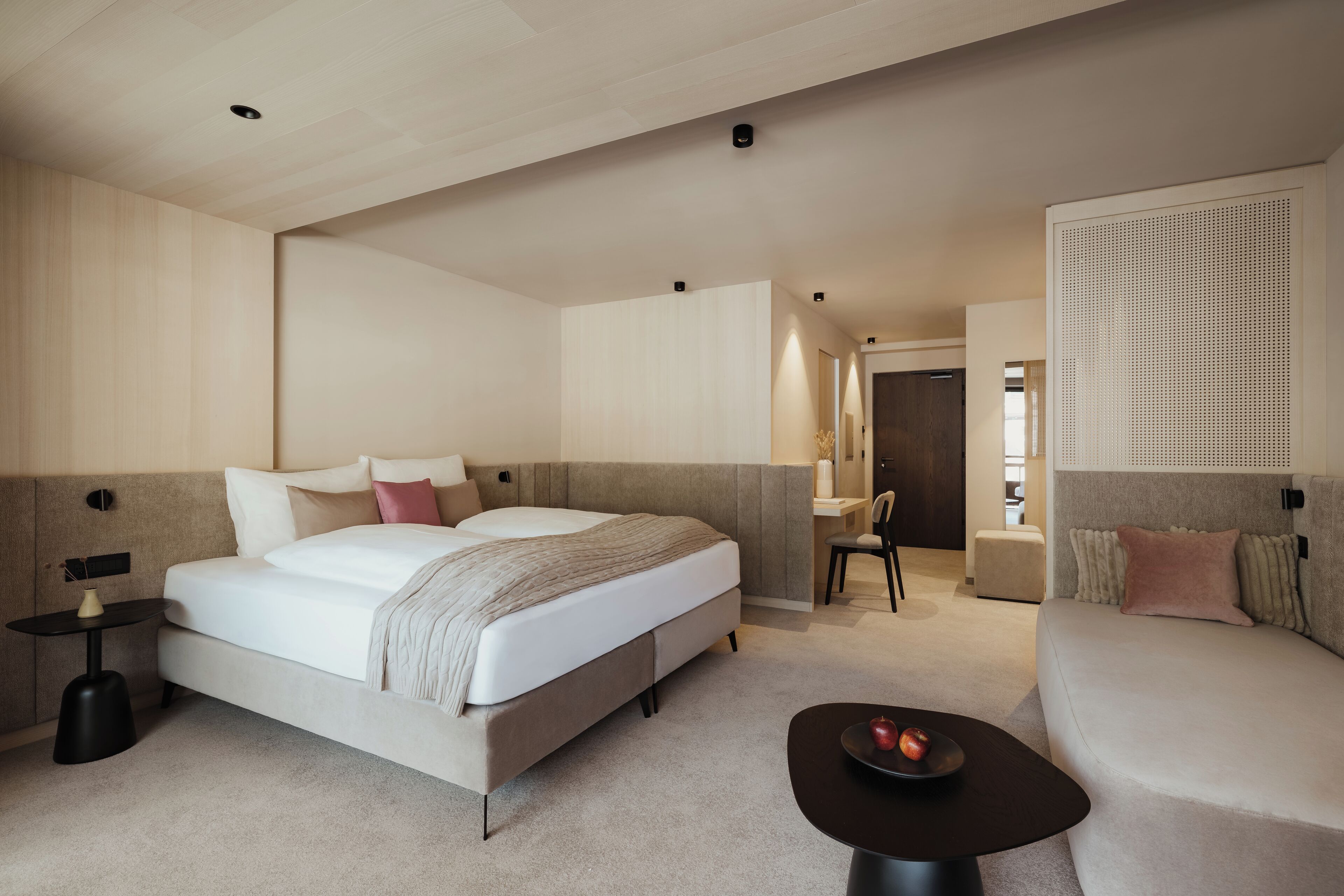 A modern hotel room with a large bed, a sofa, a small table with fruit, and a desk with a chair.